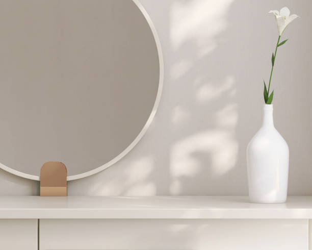 丸い化粧鏡付きのクリーム色のドレッシングテーブルのミニマルでモダンで現代的なデザイン、白い壁の寝室の窓からのまだらの日光の下での花瓶
