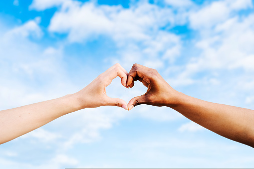 Two multiethnic hands join together to form a heart with a blue sky background. Concept of hope and love between different races, peace between humans and against hatred and racism. High quality photo