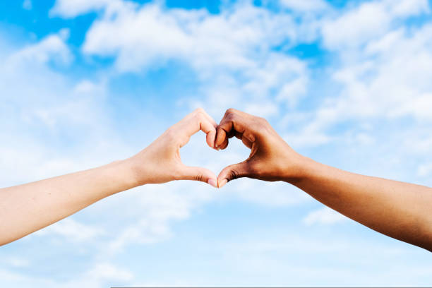 Two multiethnic hands join together to form a shape heart with a blue sky clouds background. Concept of hope and love between different races, peace between humans and against hatred and racism. Two multiethnic hands join together to form a heart with a blue sky background. Concept of hope and love between different races, peace between humans and against hatred and racism. High quality photo couple hope sky blue stock pictures, royalty-free photos & images