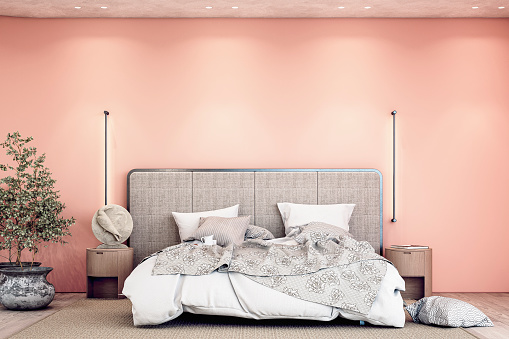 Luxury bedroom with a gray textile headboard bed, white and gray sheets - bedding,  night tables, a sculpture, pillows, a beige carpet, and vertical wall lights on the hardwood floor. A large eucalyptus plant in a pot in front of a rounded apricot pink orange empty plaster wall with copy space and built-in reflector lights on the ceiling. Front view. 3D rendered image.