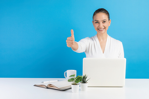 Smiling businesswoman sitting at her office desk in front of a blue wall doing thumbs up gesture in front of blue wall