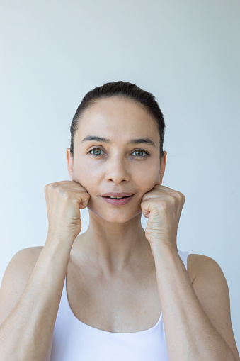 Caucasian female is doing face yoga gymnastics for non-surgical rejuvenation and is looking at camera in front of white background. Anti aging concept.