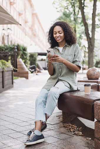 Smiling african-american student girl looking at mobile phone outdoor. People, technology, connection, mobile apps, communication and active lifestyle concept