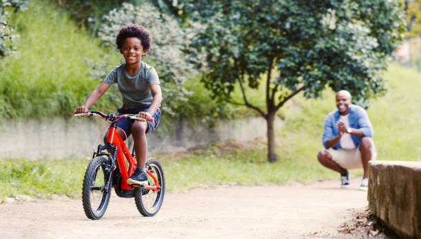 Black family, father and boy on bike in park outdoors, having fun and bonding. Love, care and man teaching child how to ride bicycle, clapping and enjoying learning time together in nature outside. stock photo