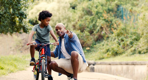istock Bicycle, learning and black man teaching kid to ride on path in garden or park, child development and growth. Nature, trust and support from dad for boy riding bike, safety and security from family. 1439733535