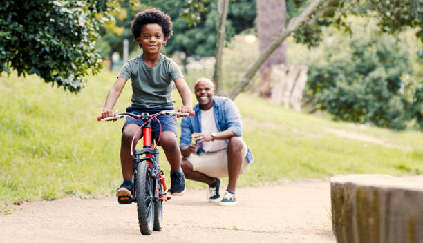 Little boy, bicycle and happy father in park for playful fun, bonding or time together in the outdoors. Proud father watching child in cycling, riding or cruising with smile on a bike in nature stock photo
