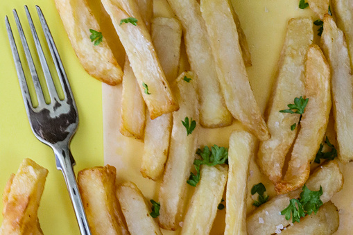 Stock photo showing close-up, elevated view of homemade pile of crispy golden French fry chips on greaseproof paper sat on yellow background besides a fork, ready to eat.