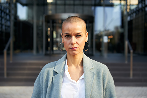 Serious middle-aged empowered woman with shaved hair looking at camera. Business people outdoors on background with buildings corporate. High quality photo