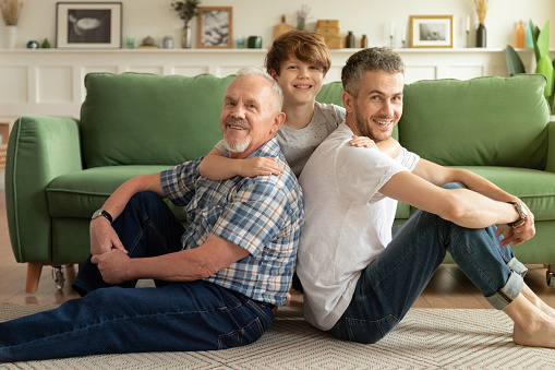Joyful multigenerational male family sitting on warm floor in living room. Happy schoolboy enjoying weekend with smiling grandpa and young handsome dad at home. Father's day and parenthood concept
