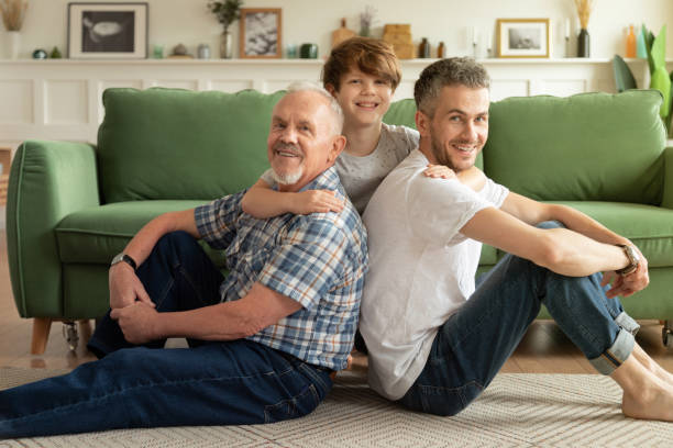 Happy multi-generational family father, son, grandfather hug each other sitting on floor in cozy home. Male generations Joyful multigenerational male family sitting on warm floor in living room. Happy schoolboy enjoying weekend with smiling grandpa and young handsome dad at home. Father's day and parenthood concept grey hair on floor stock pictures, royalty-free photos & images