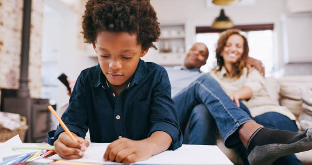 istock Child, learning and education while drawing art with colors for artwork, homework and creative activity for development. Black boy at table writing in book with parents watching their kid learn 1439756181