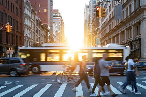 People, cars, bikes and buses traveling through a busy intersection on 5th Avenue and 23rd Street in New York City with sunlight shining in the background