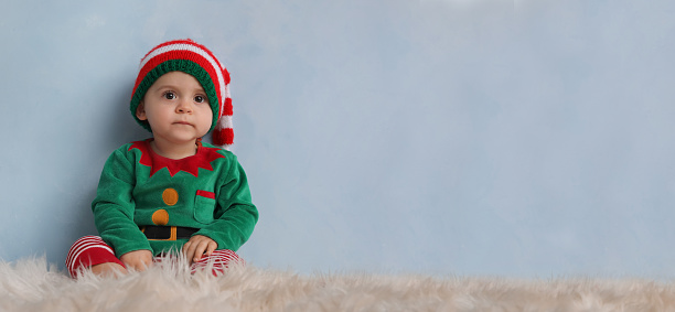 Cute baby wearing elf costume on fluffy carpet near light blue wall, banner design with space for text. Christmas celebration