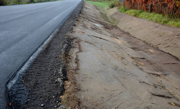 une route nouvellement construite, sur la couche de laquelle vous pouvez voir des couches successives de chape et de tapis d’asphalte. la zone environnante est inclinée dans un fossé avec une gouttière en béton. drainage des eaux pluviales - ditch photos et images de collection