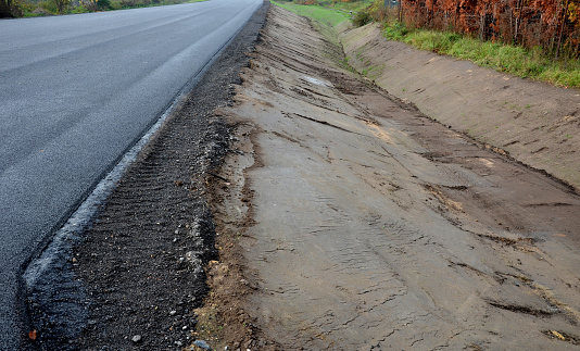 A newly built road, on the layer of which you can see successive layers of screed and asphalt carpet. the surrounding area is sloped into a ditch with a concrete gutter. rainwater drainage, tarmac