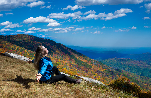 Smiling woman sitting with outstretched arms in autumn mountain scenery. Happy woman relaxing on top of the mountain. Blue Ridge Parkway, Near Asheville, North Carolina, USA.