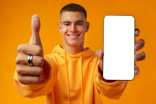 istock Smiling young man showing mobile phone blank white screen on yellow background 1439763311
