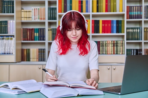 Teenage female student studying in library. Girl in headphones using laptop, looking at screen, watching webinar, making notes in notebook. Youth, technology, education, college, university concept