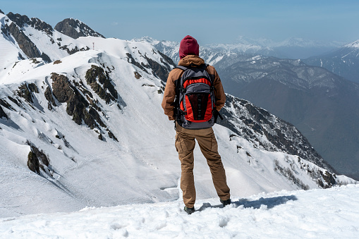 rear view of young man hiker with backpack looking at view of snowy peaks of mountains in winter against blue sky hiking copy space enjoying nature