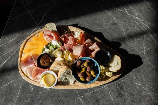 A directly above shot of a French styled food platter on a wooden board which includes sliced bread, olives, salami, parma ham, chutney, selection of cheeses and mortadella.