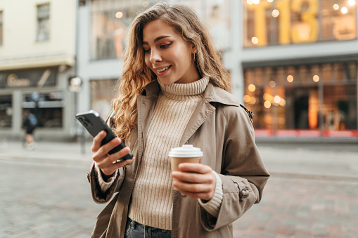Young woman wearing autumn coat walking with smartphone and coffee cup in a city street, portrait