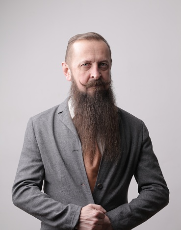 A vertical shot of a man with a long beard and a mustache standing in front of a grey wall