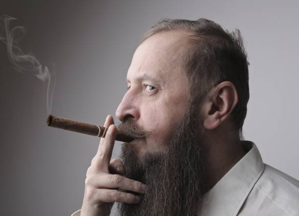 istock Man with a long beard and a mustache smoking a cigar with a grey wall on the background 1439777876