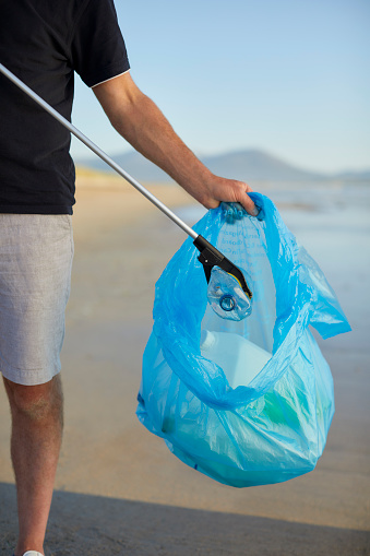 Midsection of man putting water bottle in plastic bag with litter picker. Male activist is cleaning beach. He is an environmentalist.
