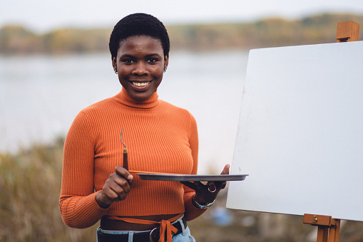 A young African female adult posing for portrait photos with her painting equipment outdoors, by the river.