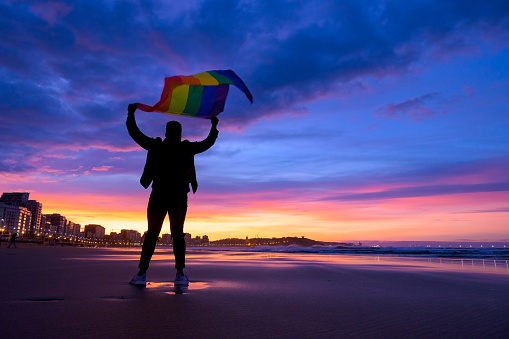 An amazing shot of a young male standing on a beach and holding in hands a gay flag