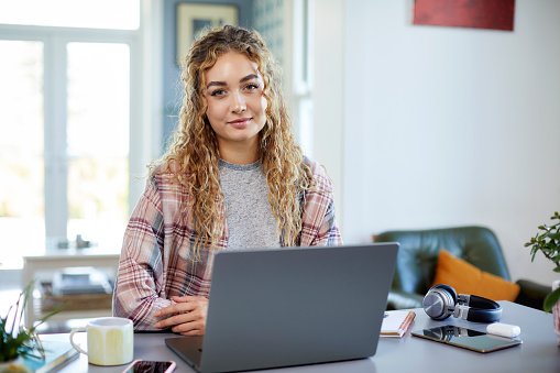 Portrait of young female entrepreneur sitting with laptop on table at home. Beautiful female professional has blond curly hair. She is telecommuting from apartment.