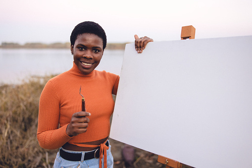 A young African female adult posing for portrait photos with her painting equipment outdoors, by the river.
