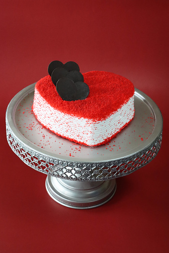 Stock photo showing elevated view of cake stand containing a whole, heart-shaped red velvet cake covered in butter cream and decorated with dehydrated raspberry powder and three chocolate hearts, displayed against a red background.