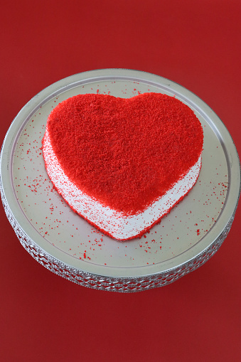 Stock photo showing elevated view of cake stand containing a whole, heart-shaped red velvet cake covered in butter cream and decorated with dehydrated raspberry powder, displayed against a red background.