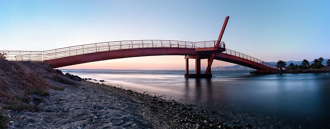 A Horizontal panoramic shot of a red metal bridge crossing over the sea. Long exposure photography of a seashore landscape at the sunset.