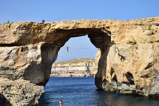 Dwejra, Gozo, Malta – October 11, 2014: A man cliff diving, jumping off the Azure Window into the sea below, at Dwejra Gozo. A natural arch formed by sea erosion of the cliffs in Malta