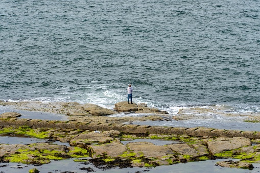 Whitley B, United Kingdom – July 21, 2022: A man stands on a rock while sea fishing, in Whitley Bay, UK.