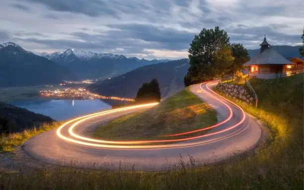 The light trails of cars on a road to the water and illuminated building surrounded by mountains in Zell am See, Austria