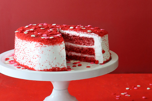 Stock photo showing close-up view of cake stand containing a cut, heart-shaped red velvet cake covered in butter cream and decorated with dehydrated raspberry powder and sugar sprinkle hearts, displayed against a red background.