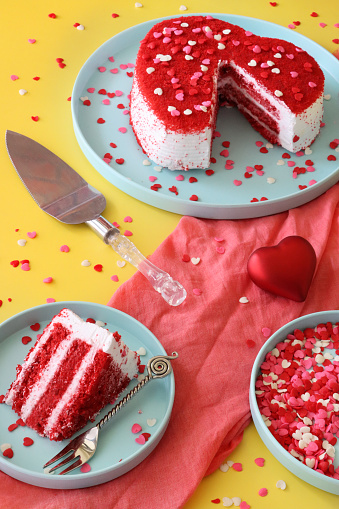 Stock photo showing elevated view of plate containing a cut, heart-shaped red velvet cake covered in butter cream and decorated with dehydrated raspberry powder and sugar sprinkle hearts, displayed against a yellow background.
