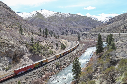 Floriston, California, United States – April 12, 2020: A freight train passes along the Truckee River in a canyon beneath the peaks of the Sierra Nevada mountains.