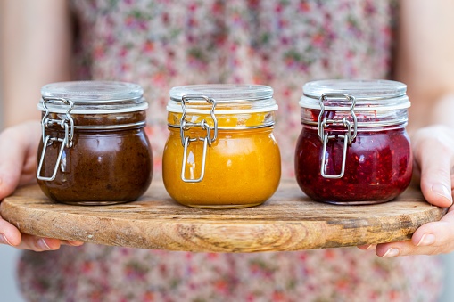 A closeup shot of a woman holding a wooden plate with plum, apricot, raspberry jam glass jars