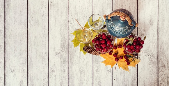 On wooden table are vases with ripe cherries, jug of wine, glass of mineral water, yellow wedge leaves and fir cones. View from above.