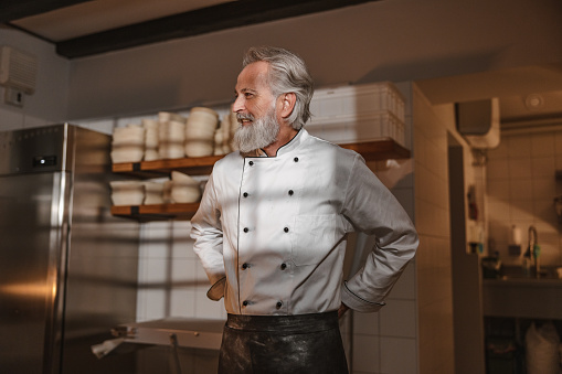 Three-quarter side shot with blurred background of an experienced Caucasian mature baker looking away and wearing a white chef's uniform and tying a black apron as a part of his uniform and routine before starting his early morning shift at the bakery surrounded by all kitchen supplies.