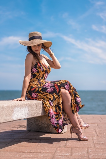 A vertical shot of a female in floral dress and hat captured by the ocean in San Sebastian, Spain