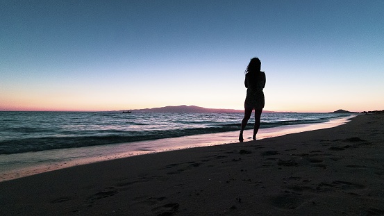 A silhouette of a lonely female walking on the beach near the sea during sunset