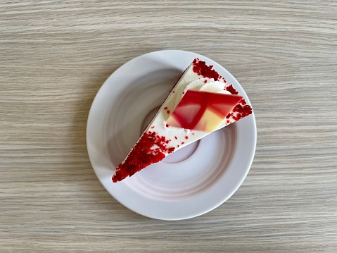 A top view of a piece of red velvet cake decorated with strip of white chocolate on a white plate lying on a wooden table.
