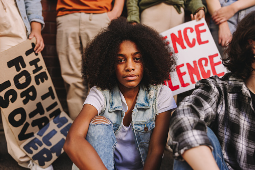 Black teenage girl looking at the camera while sitting with a group of youth peace activists. Multiethnic young people displaying posters and banners while protesting against racial violence.