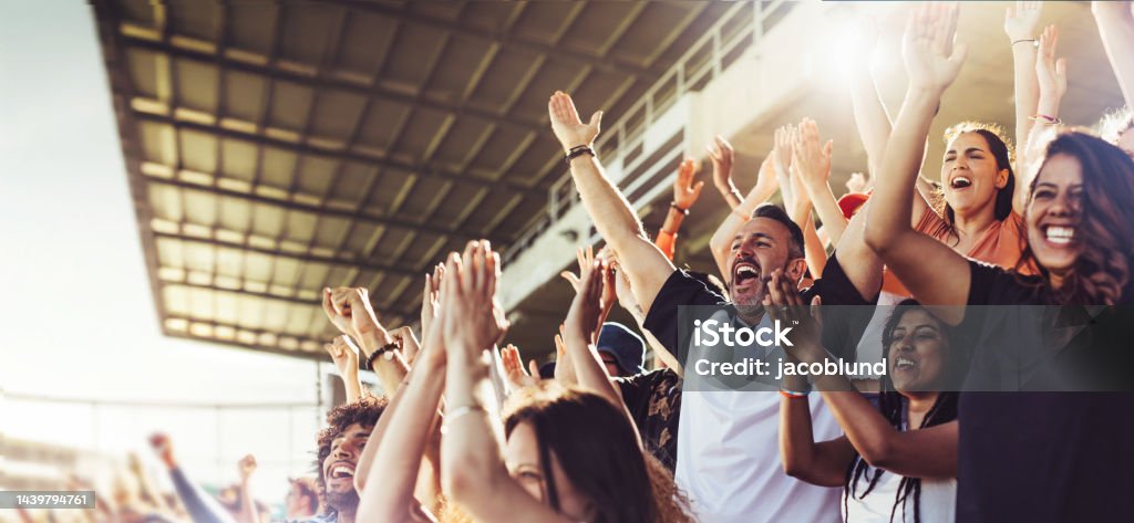 Crowd of sports fans cheering during a match in a stadium - people excited cheering for their favorite sports team to win the game Crowd of sports fans cheering during a match in stadium. Excited people standing with their arms raised, clapping, and yelling to encourage their team to win. Fan - Enthusiast Stock Photo