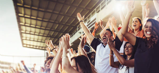 Crowd of sports fans cheering during a match in a stadium - people excited cheering for their favorite sports team to win the game Crowd of sports fans cheering during a match in stadium. Excited people standing with their arms raised, clapping, and yelling to encourage their team to win. football fans cheering stock pictures, royalty-free photos & images